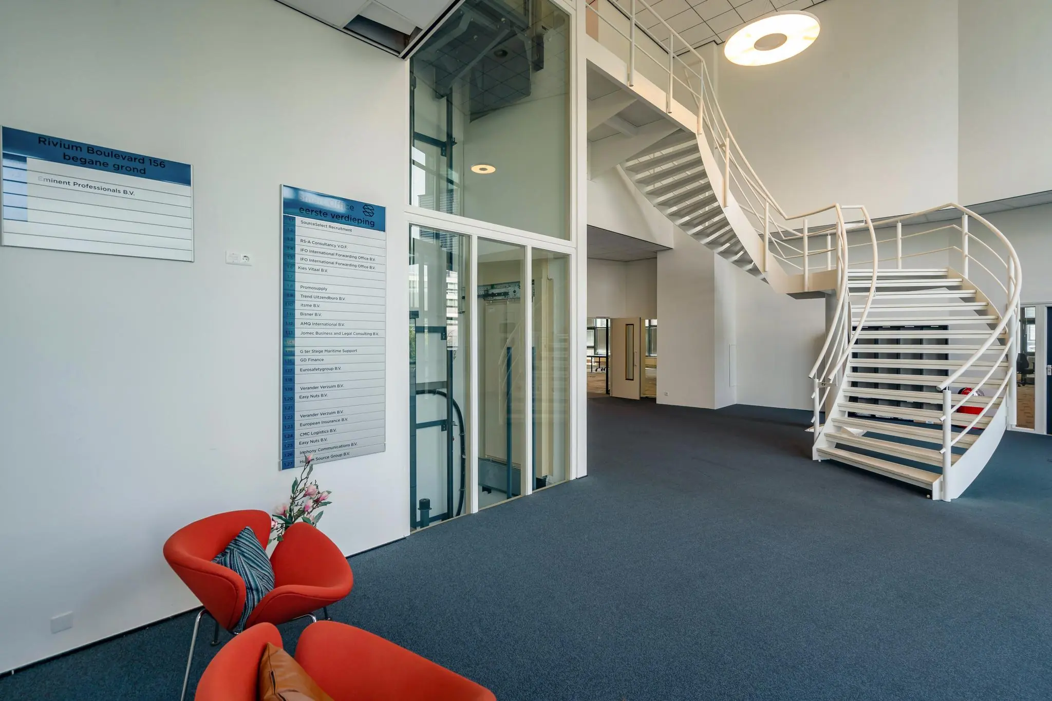 Moderne bedrijfsentree aan de Rivium Boulevard 156 met een witte wenteltrap, lift en informatiebord met bedrijfsnamen.